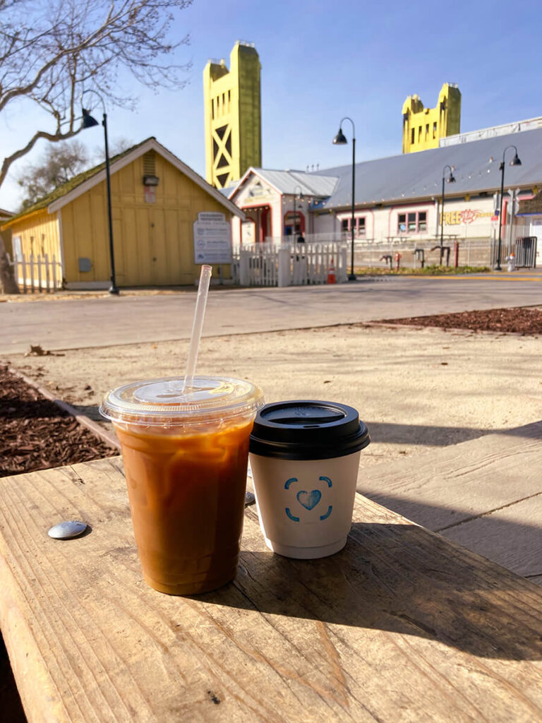 Two cups of coffee on a wooden bench in Old Sacramento. On the left is an iced coffee in a plastic cup and straw, with a long shadow cast in front of it. On the right is a hot coffee with a blue heart on it. At a distance, you can see buildings in Old Sacramento and the Tower Bridge that connects Old Sacramento and West Sacramento. This was taken on a sunny February morning, with trees with no leaves in the background.