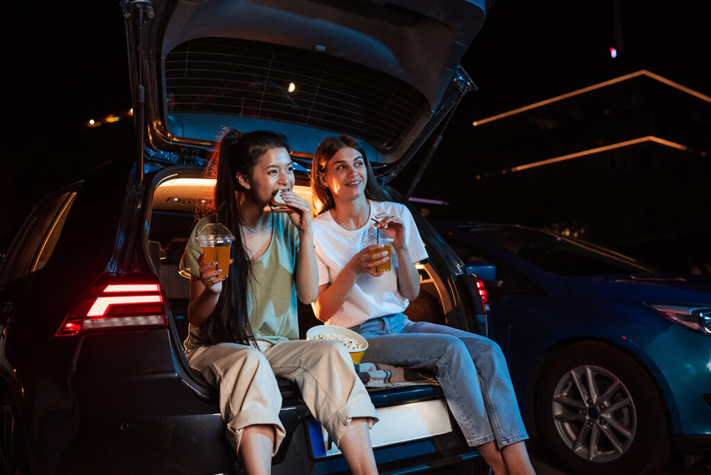 two women sitting in the back of a car eating food watching a drive-in movie at night