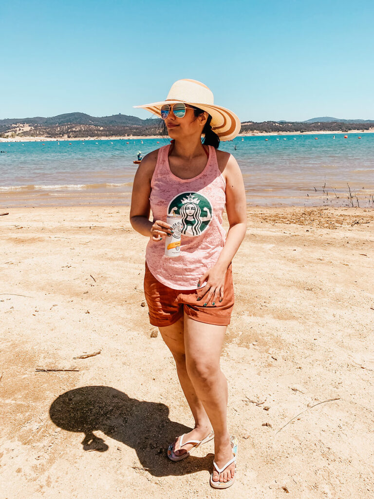 Hannah drinking a white claw standing in front of Folsom Lake on a hot, sunny, day. Folsom Lake has sand and calm waters. At a distance, there are some people kayaking.