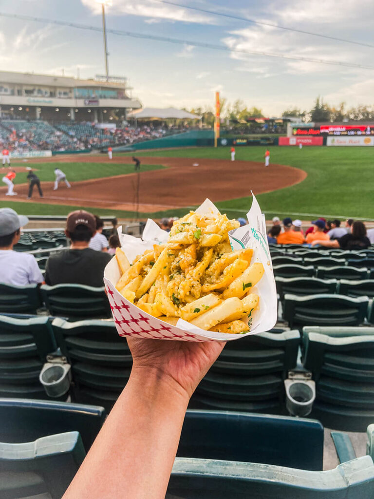 Hannah holding a paper tray of garlic fries at Sutter Health Park in West Sacramento, CA. Beyond the distance are the Sacramento River Cats playing against another team. This photo was taken during sunset