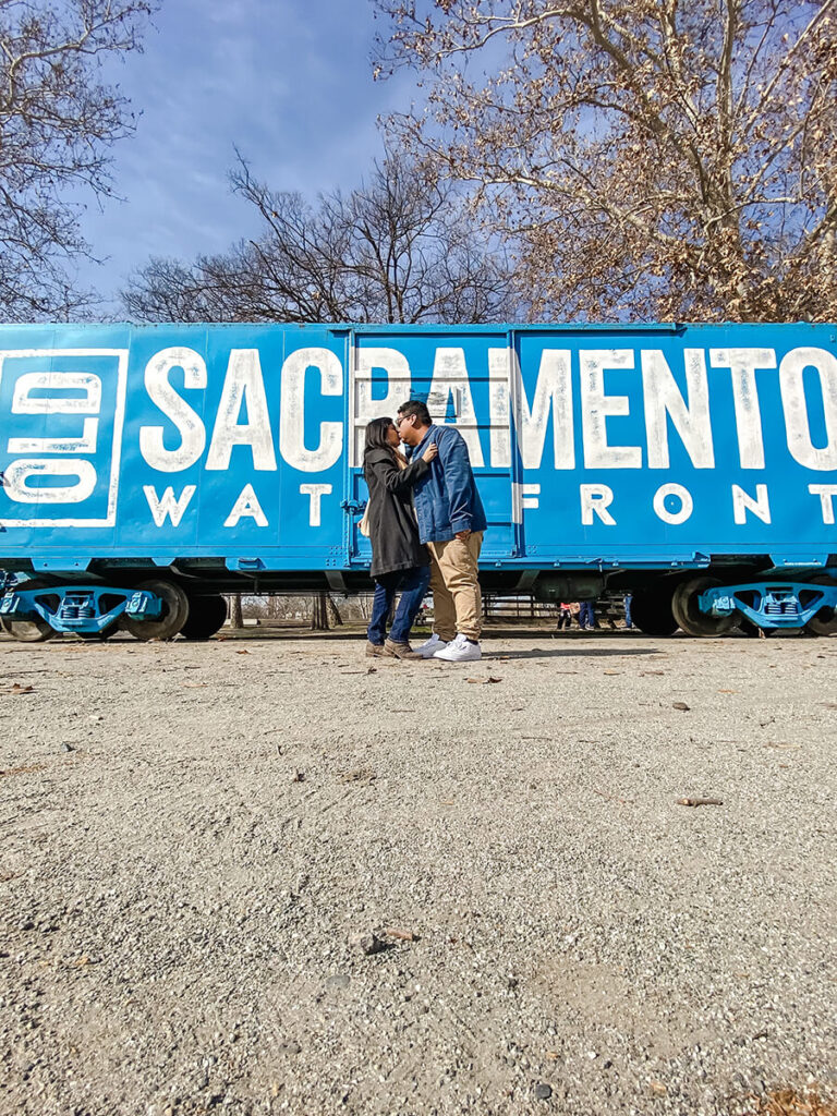 Hannah and Cecilio kissing in front of the large blue Old Sacramento Waterfront sign in Sacramento, CA on a sunny day during winter. Behind them are some trees with dead leaves.