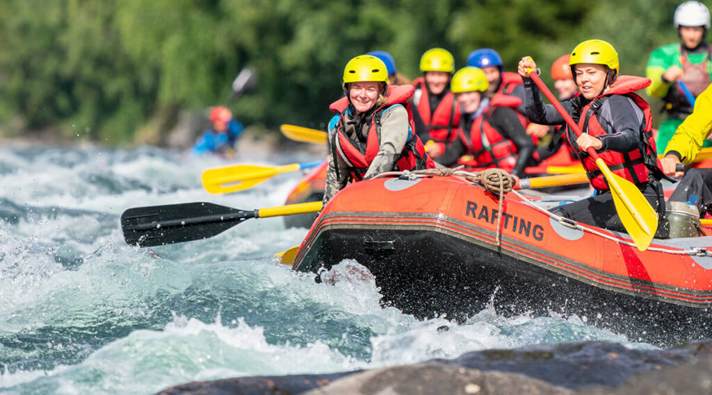 A group of people white water rafting down the river. They are wearing red life jackets, holding yellow and red paddles, and blue and yellow helmets. They are on an orange rafting boat 