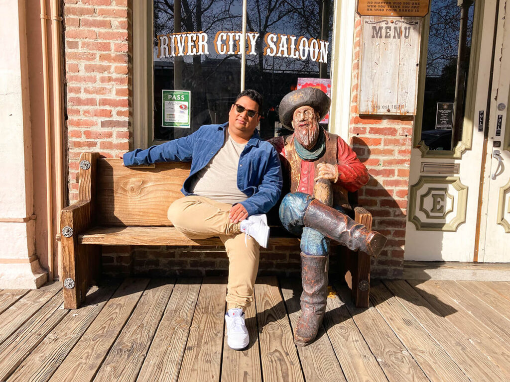 Cecilio sitting on a wooden bench with a cowboy statue in front of the River City Saloon in Old Sacramento. Cecilio is mimicking the statue's pose by crossing his leg.